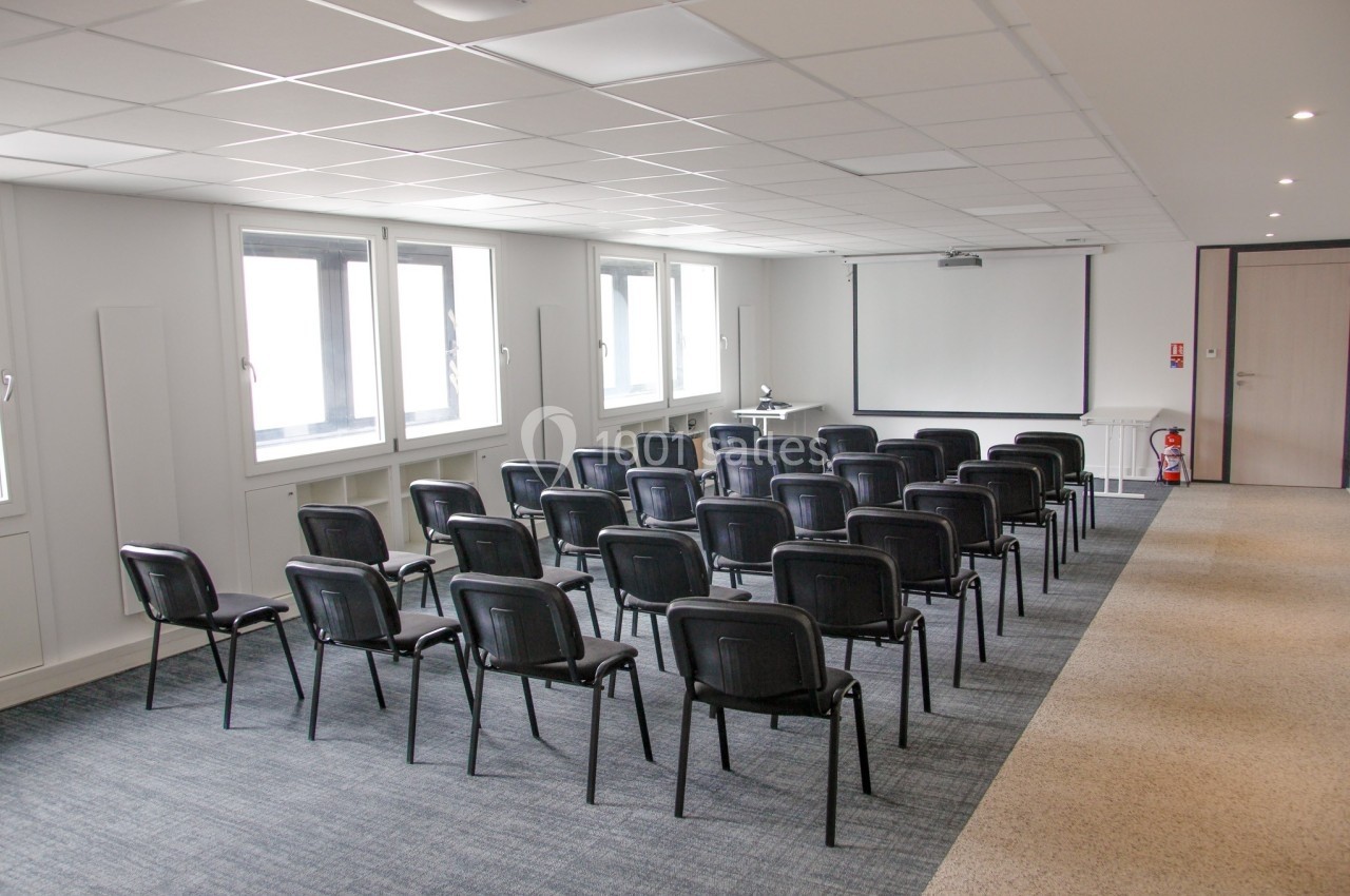 Salle de réunion lumineuse avec rangées de chaises noires, écran de projection et fenêtres sur un mur latéral.