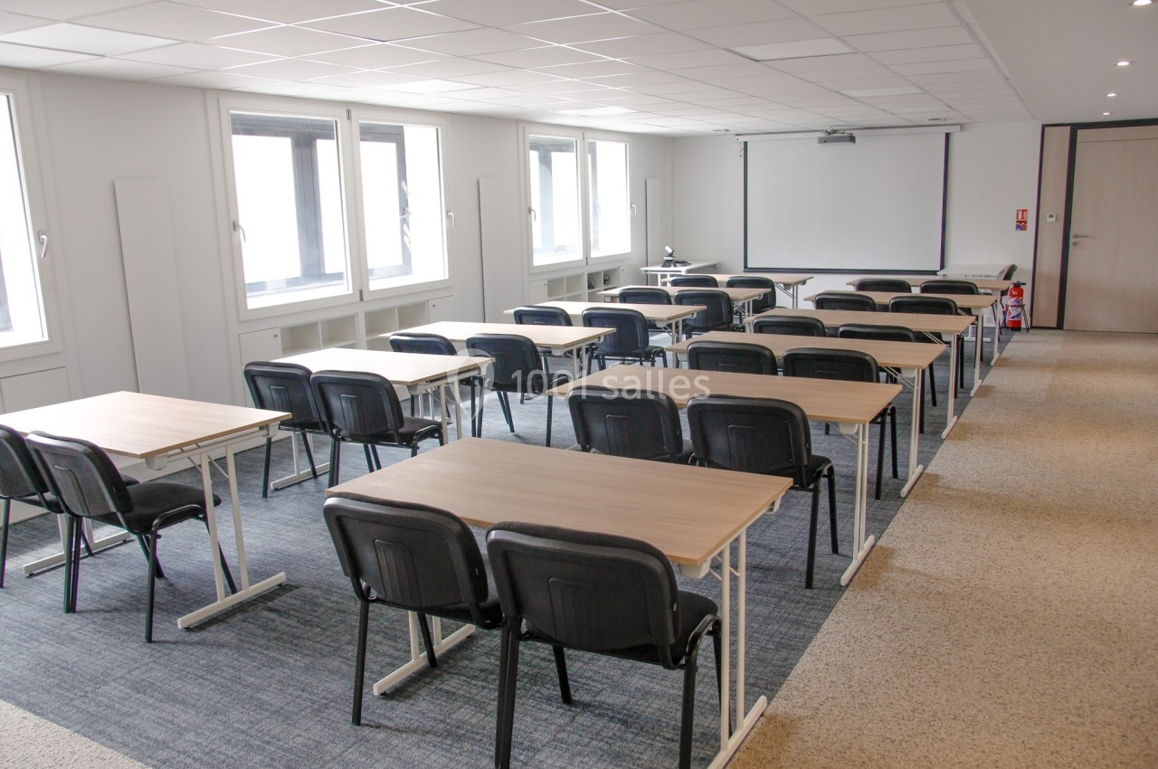 Salle de formation lumineuse avec des tables, chaises noires et un écran de projection au fond.