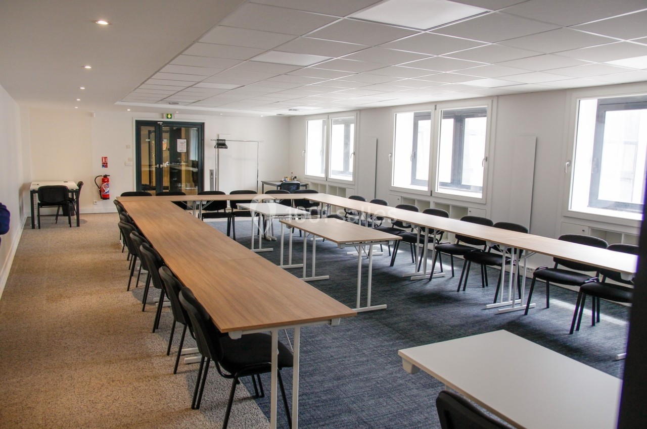 Salle de réunion lumineuse avec des tables disposées en U, des chaises noires et un sol en moquette grise.