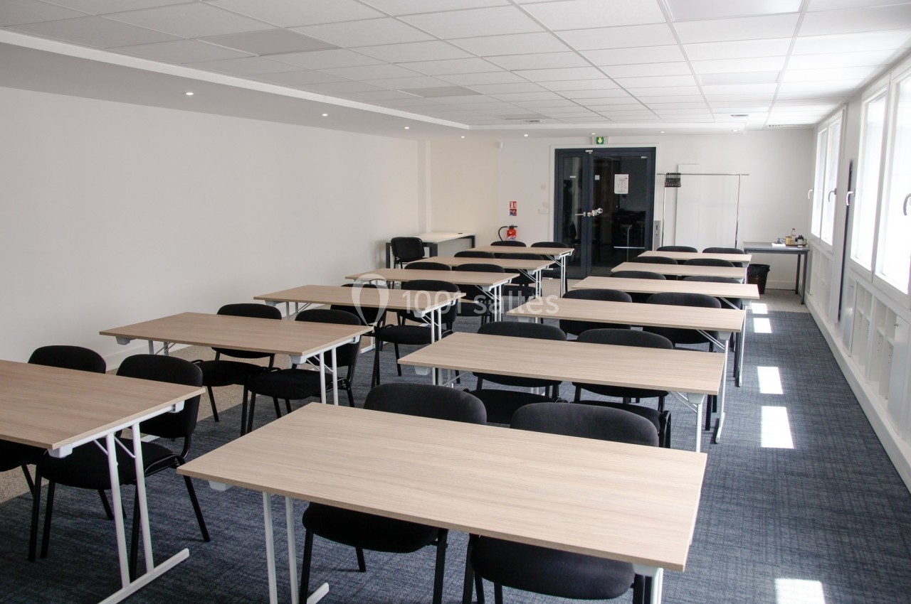 Salle de réunion lumineuse avec plusieurs tables en bois clair et chaises noires, disposées en rangées.