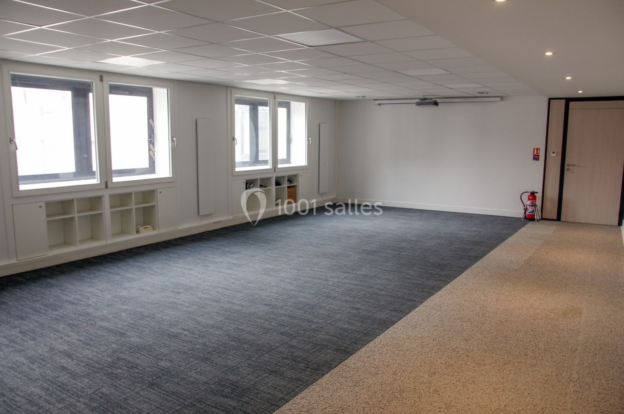 Salle vide avec moquette grise et beige, murs blancs, fenêtres vitrées, et étagères intégrées sous les fenêtres.