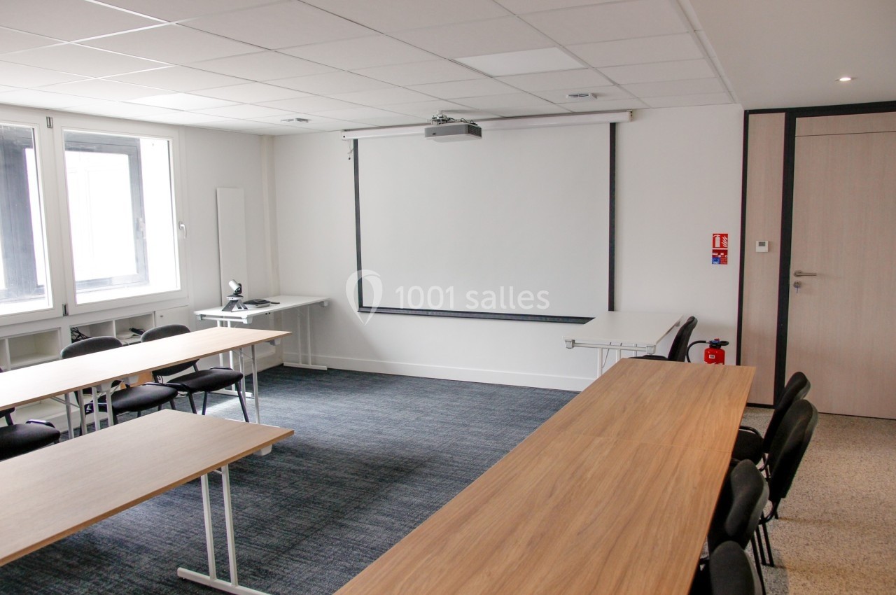 Salle de réunion lumineuse avec tables en bois, chaises noires, écran de projection et fenêtres.