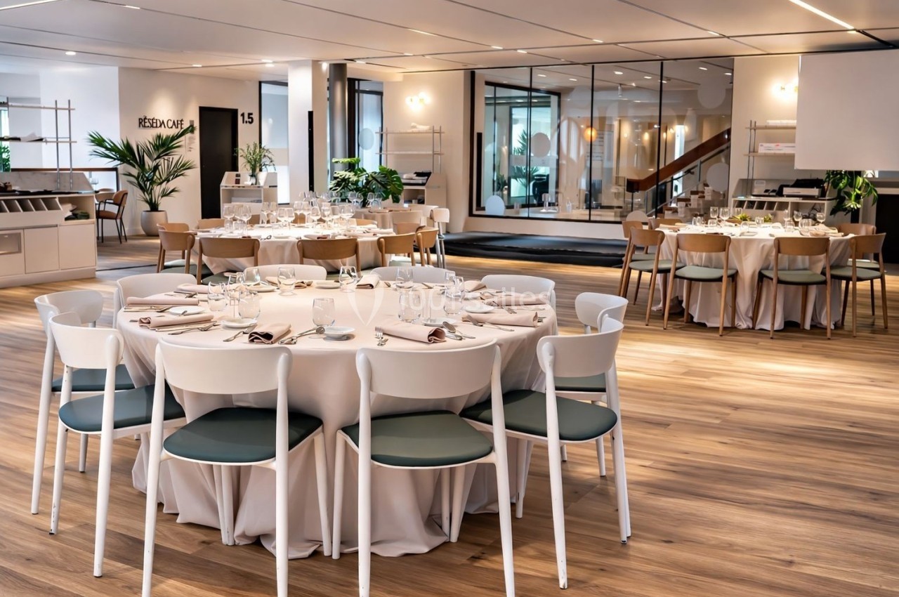 Salle lumineuse aménagée avec des tables rondes dressées pour un repas, entourées de chaises modernes.