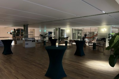 Salle spacieuse avec parquet clair, tables hautes recouvertes de nappes vertes et grandes baies vitrées en arrière-plan.