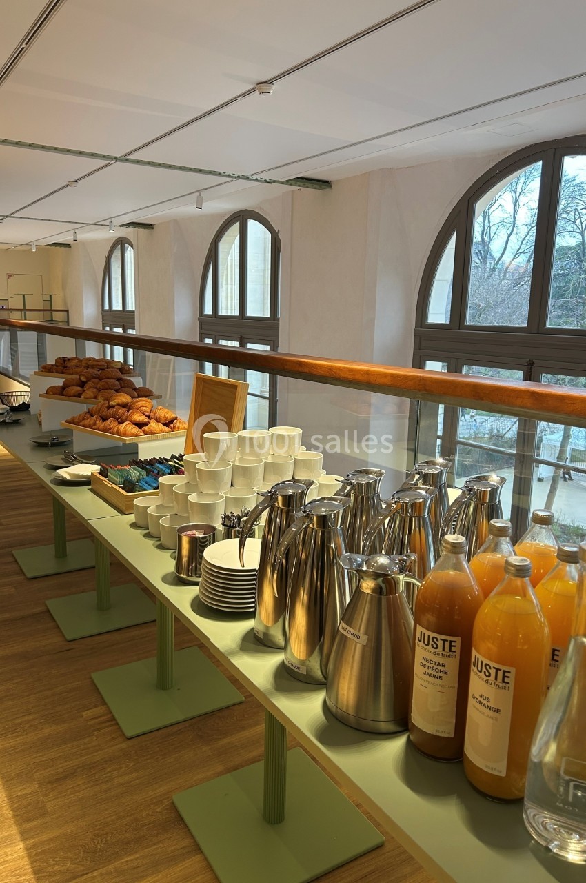 Buffet avec viennoiseries, jus d'orange, carafes en inox et tasses, disposé dans une salle lumineuse avec grandes fenêtres.