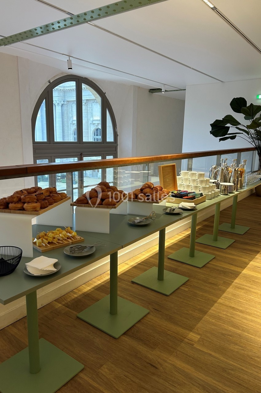Table avec viennoiseries, fruits, boissons et vaisselle, disposée dans une salle lumineuse avec une grande fenêtre en…
