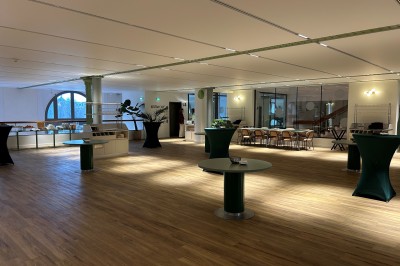 Salle spacieuse avec parquet clair, tables hautes recouvertes de nappes vertes et grandes baies vitrées en arrière-plan.