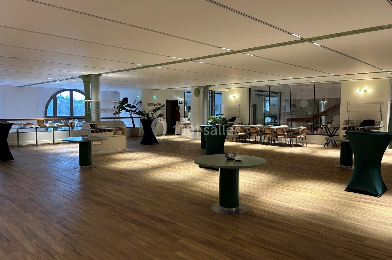 Salle spacieuse avec parquet clair, tables hautes recouvertes de nappes vertes et grandes baies vitrées en arrière-plan.