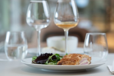 Salle élégante avec tables rondes dressées, nappes beiges, chaises blanches et décoration florale, éclairée par des baies…