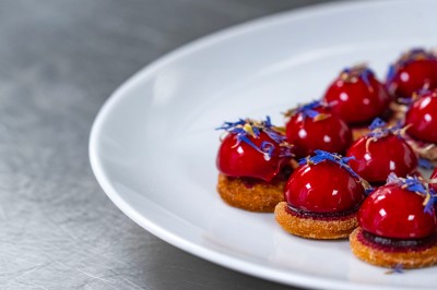 Salle élégante avec tables rondes dressées, nappes beiges, chaises blanches et décoration florale, éclairée par des baies…