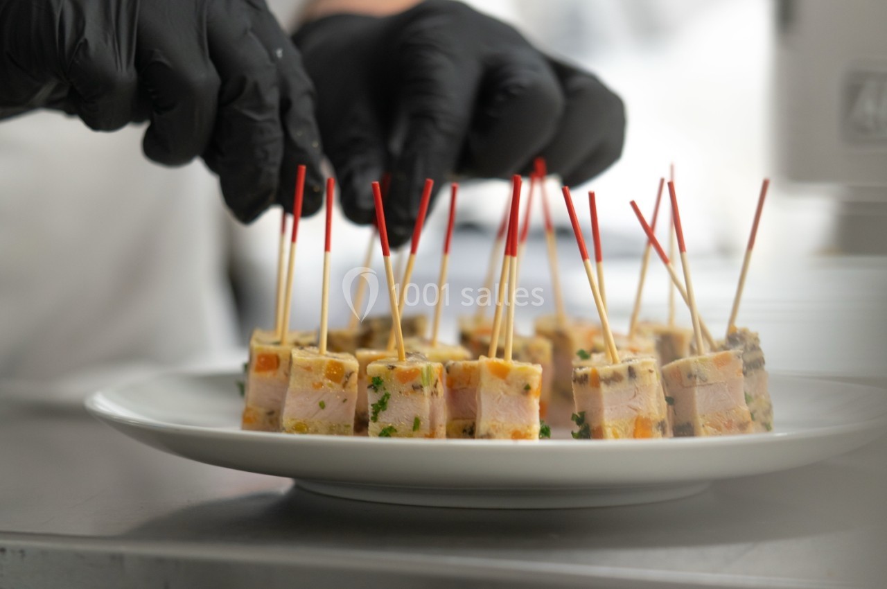 Mains en gants noirs disposant des bouchées apéritives sur des piques rouges dans une assiette blanche.