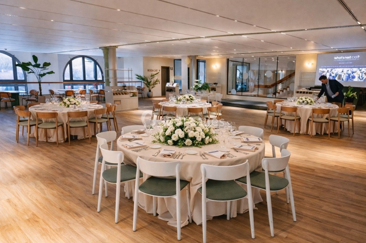 Salle élégante avec tables rondes dressées, nappes beiges, chaises blanches et décoration florale, éclairée par des baies…