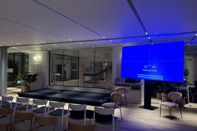 Salle élégante avec tables rondes dressées, nappes beiges, chaises blanches et décoration florale, éclairée par des baies…