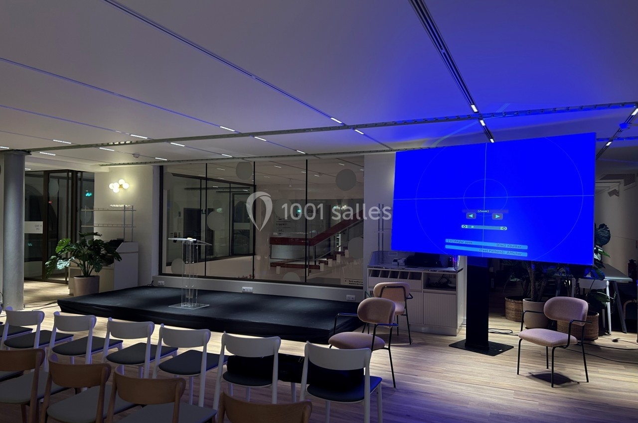 Salle de conférence moderne avec un écran géant allumé, des chaises disposées et un éclairage bleu tamisé.