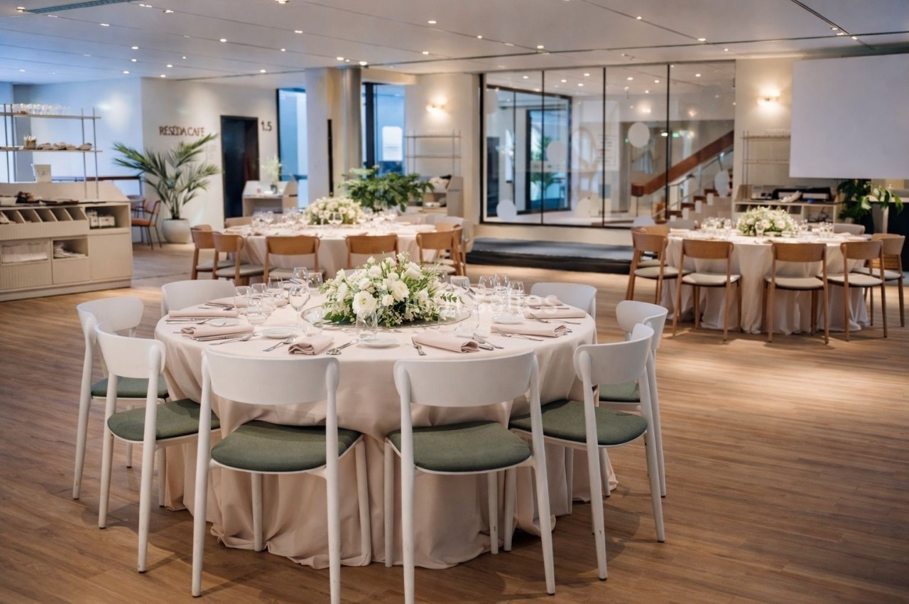 Salle de réception élégante avec tables rondes dressées, décorées de centres de table floraux et chaises blanches.