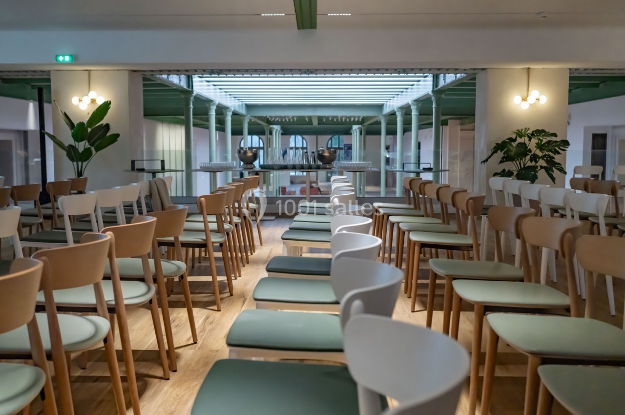 Salle lumineuse avec des rangées de chaises en bois et assises vertes ou blanches, entourée de plantes et éclairée par un…