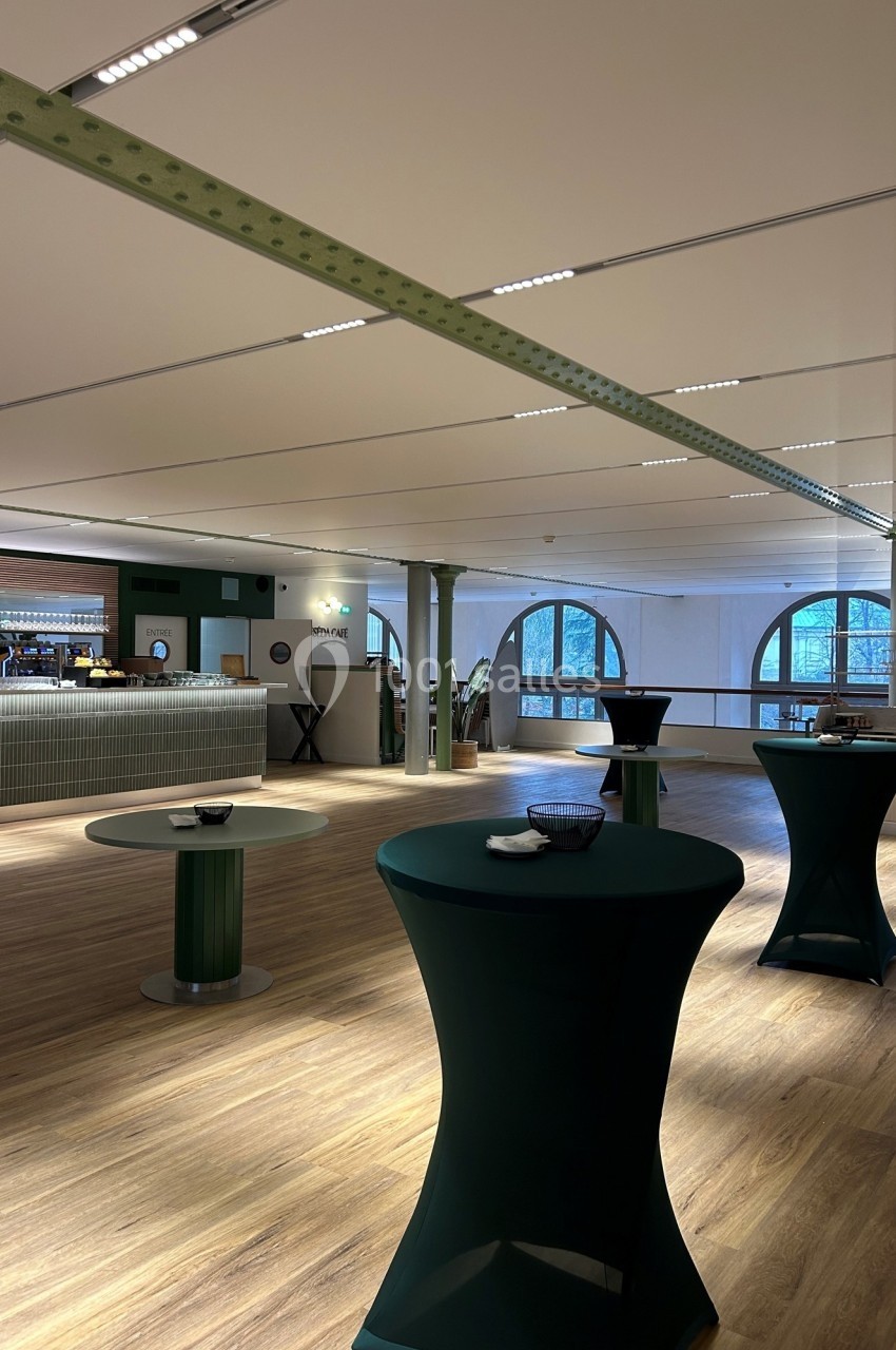 Salle lumineuse avec parquet, tables hautes drapées de vert et un bar en arrière-plan.