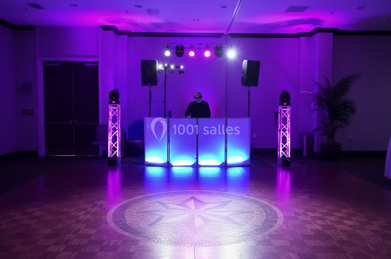 DJ mixant derrière une table éclairée par des lumières violettes et bleues, dans une salle avec piste de danse vide.