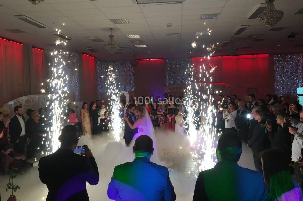 Un couple danse au centre d'une salle de réception entouré de fumée et de jets d'étincelles, devant des invités.