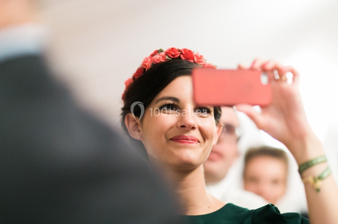Femme souriante portant une couronne de fleurs rouges, prenant une photo avec un smartphone rose.