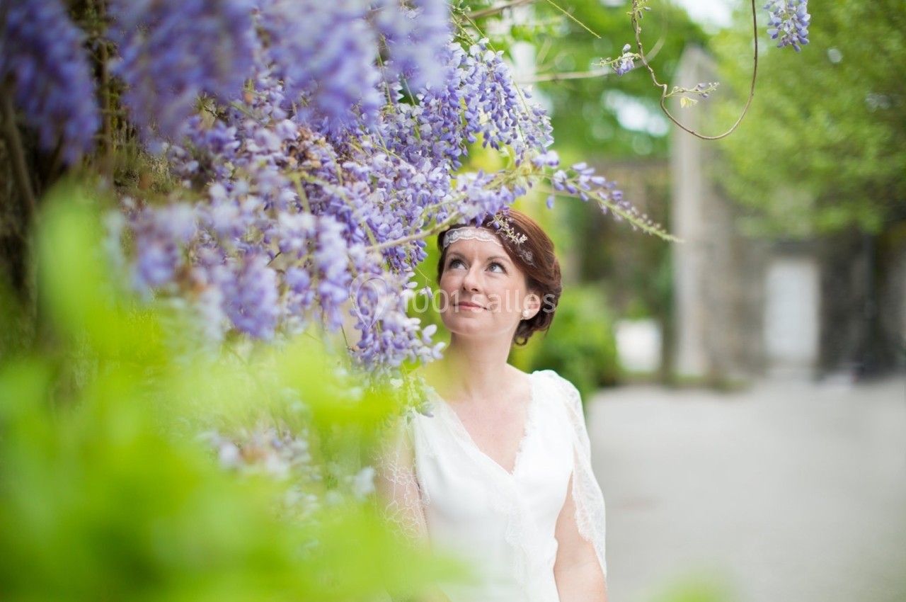 Femme en robe blanche regardant des glycines violettes en fleurs dans un jardin verdoyant.
