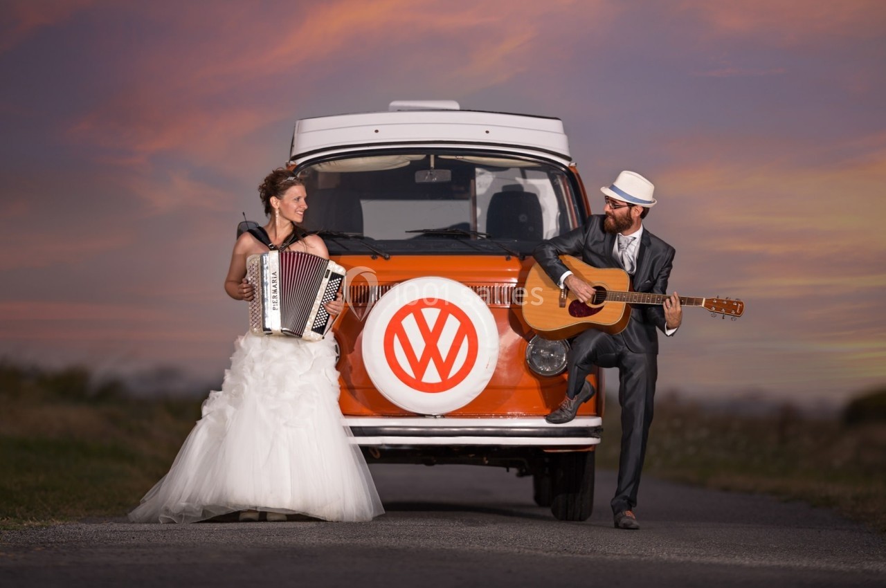 Un couple en tenue de mariage joue de l'accordéon et de la guitare devant un van orange au coucher du soleil.