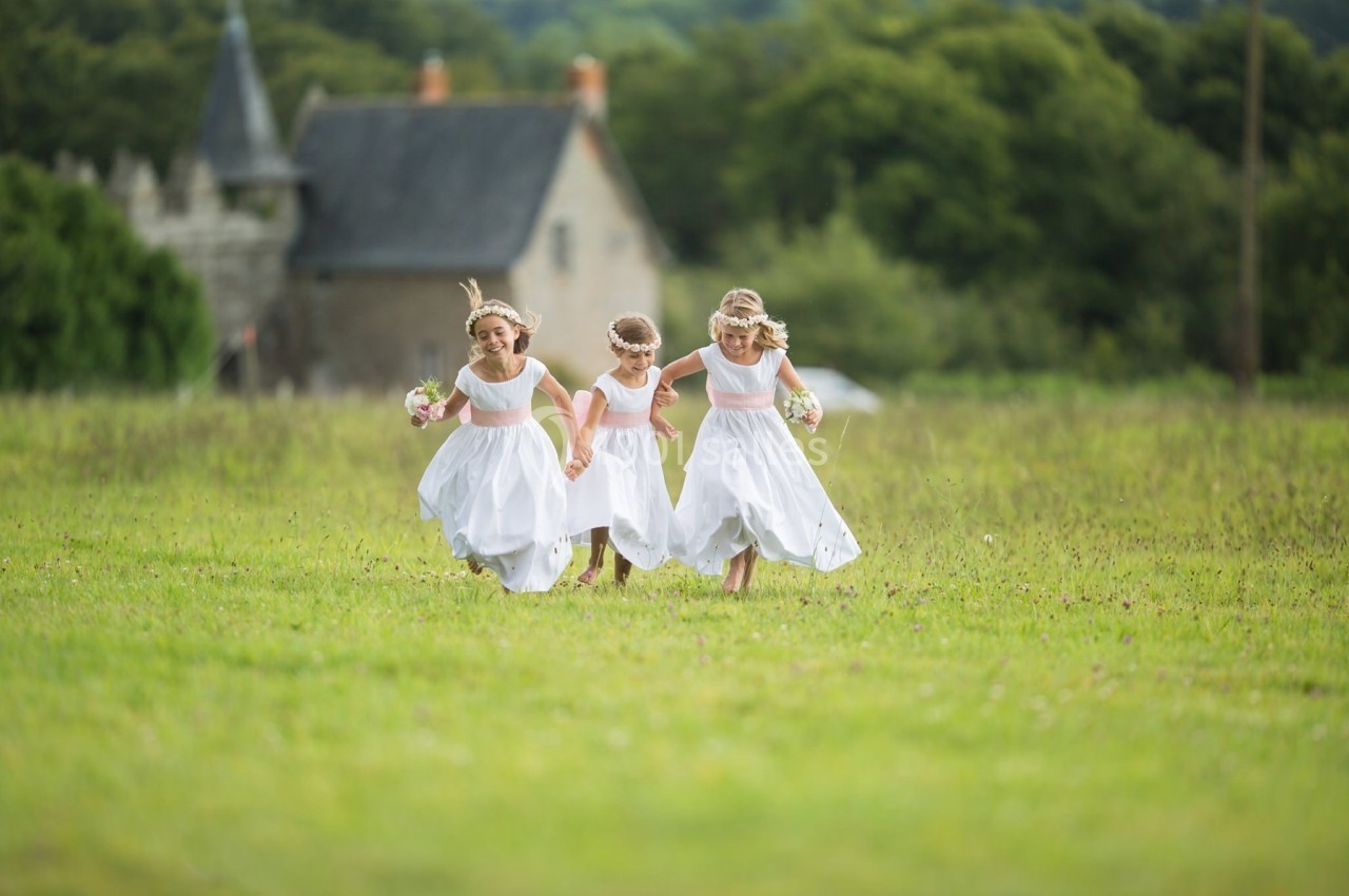 Trois jeunes filles en robes blanches courent joyeusement dans un champ verdoyant devant un bâtiment en pierre.