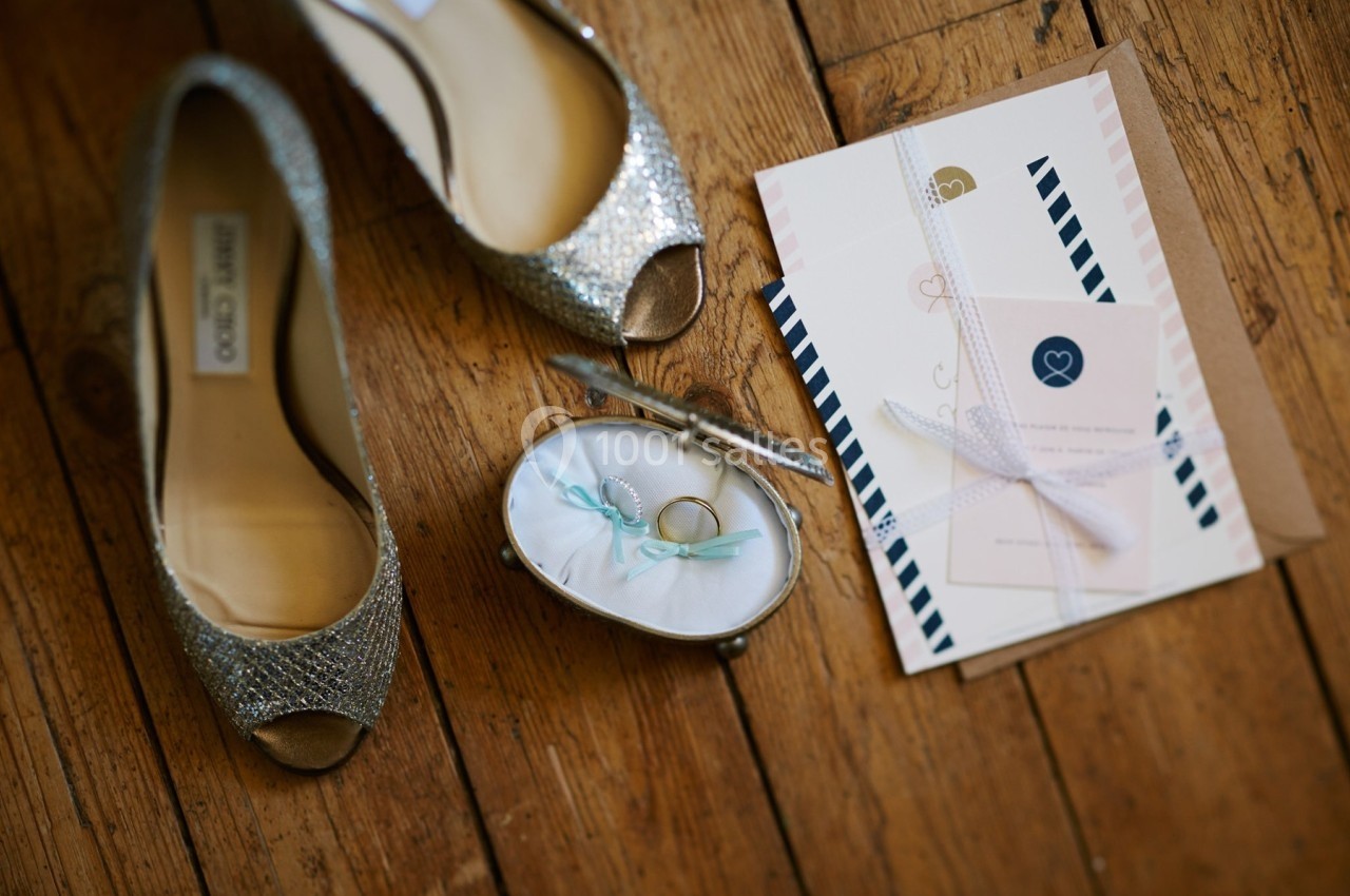 Chaussures argentées, alliances dans un écrin et faire-part de mariage posés sur un parquet en bois.