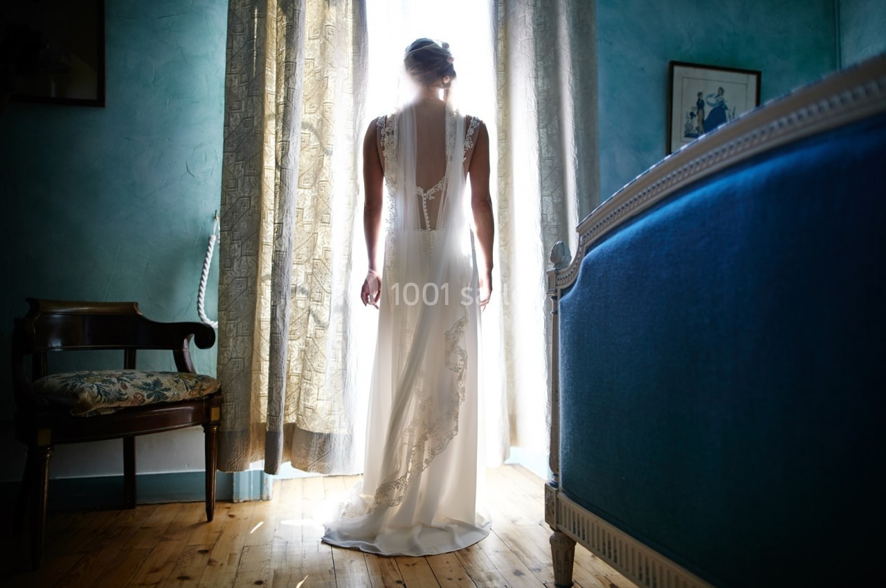 Femme en robe de mariée debout devant une fenêtre, éclairée par la lumière naturelle dans une pièce au décor classique.