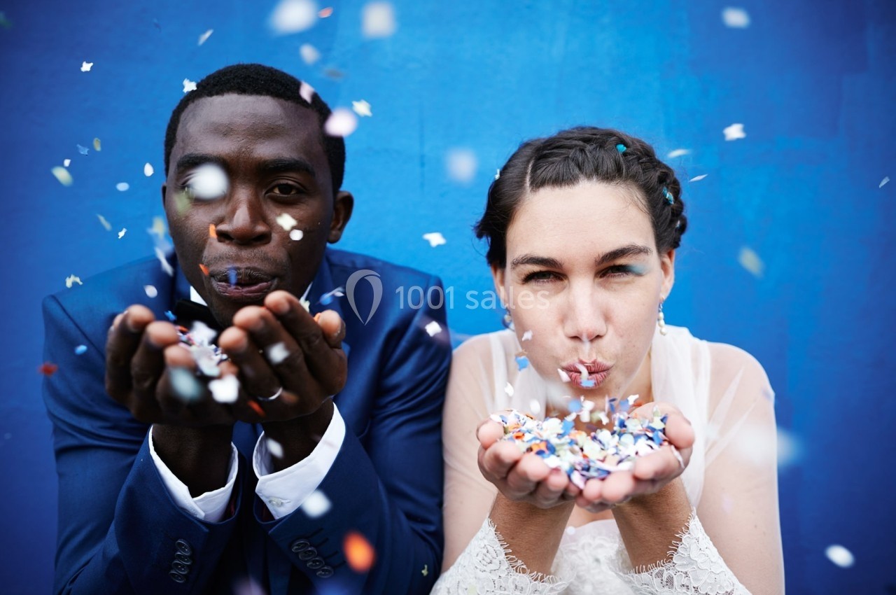 Deux personnes soufflent des confettis colorés devant un mur bleu.