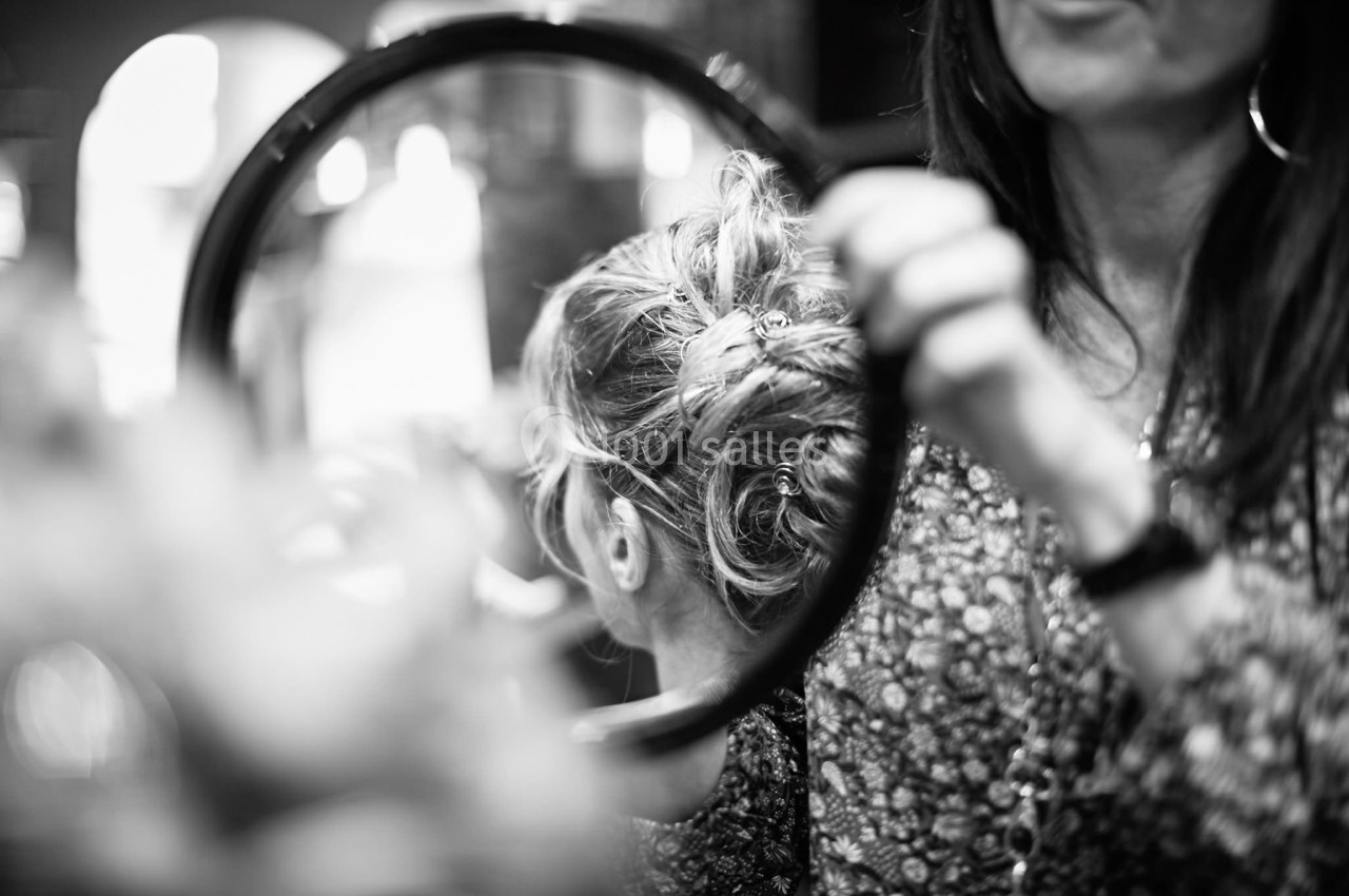 Une coiffeuse tient un miroir rond reflétant une coiffure élaborée sur une femme de dos.