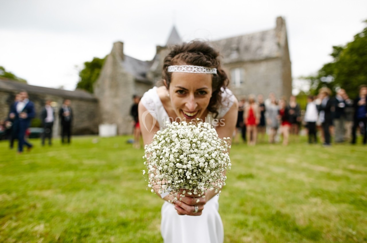Une mariée souriante tient un bouquet de gypsophiles dans un jardin devant un bâtiment en pierre.