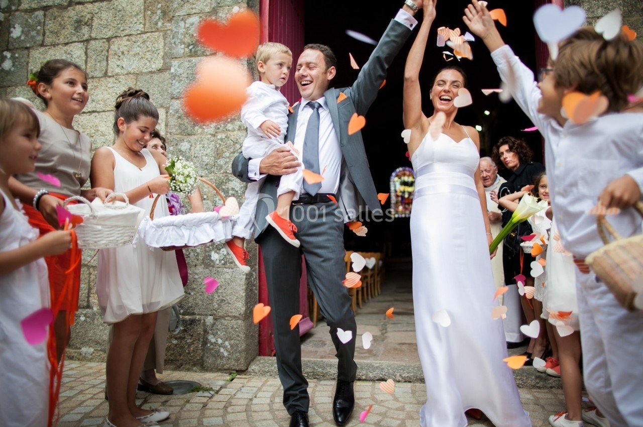 Un couple de mariés souriants sort d'une église, entouré d'enfants et de confettis en forme de cœurs.