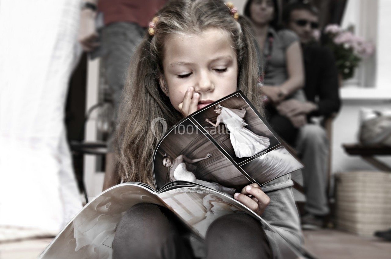 Une petite fille feuillette un magazine de photos de mariage, assise à l'extérieur devant des adultes en arrière-plan.