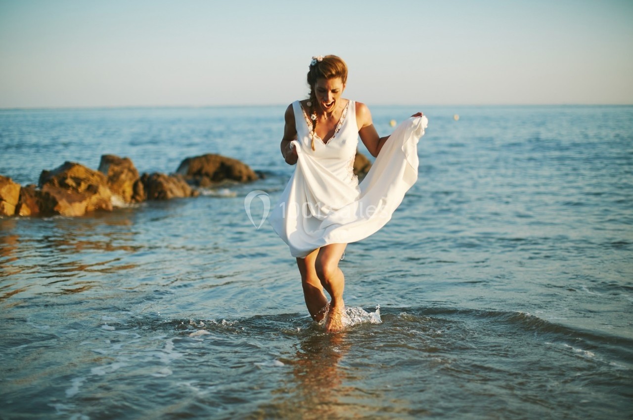 Femme en robe blanche marchant dans l'eau près de rochers, sous un ciel dégagé au bord de la mer.