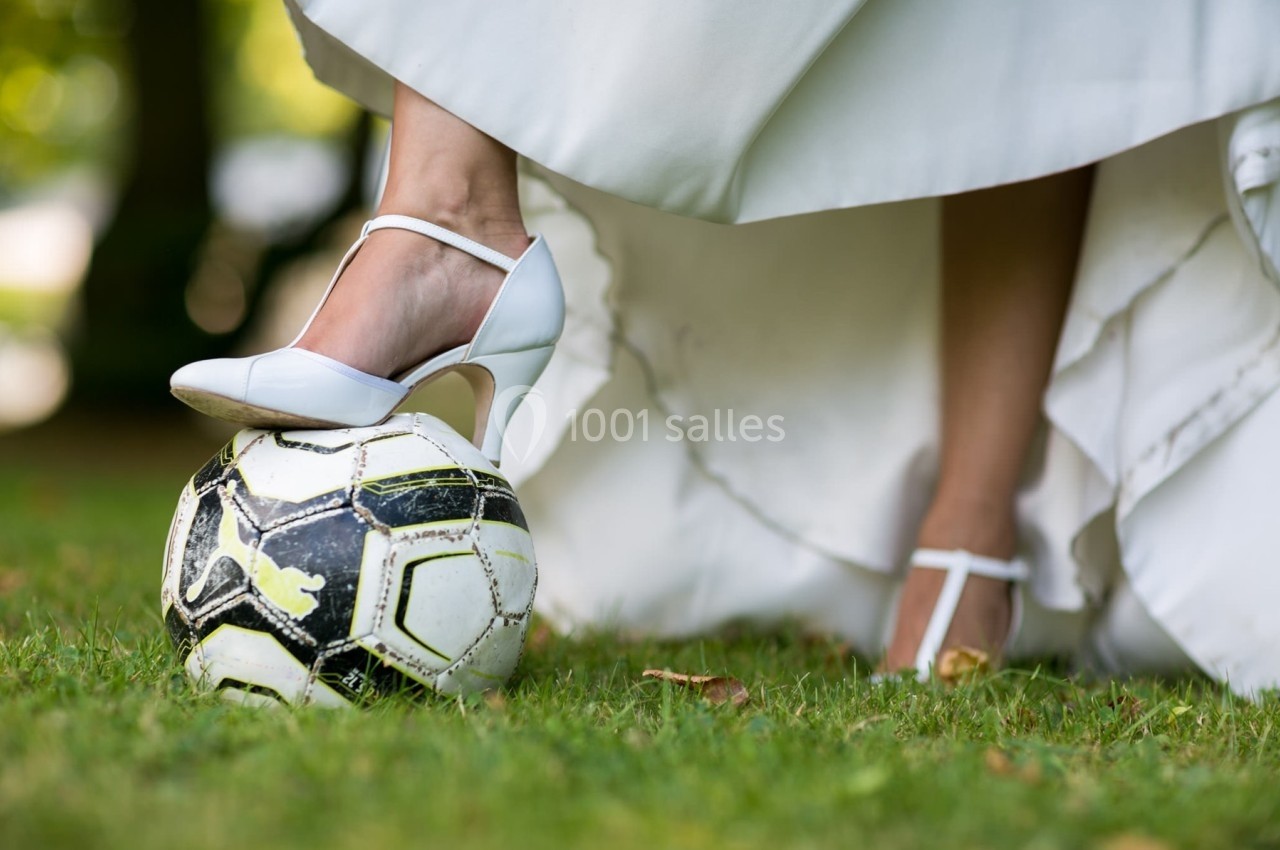 Une femme en robe blanche et talons pose son pied sur un ballon de football sur une pelouse.