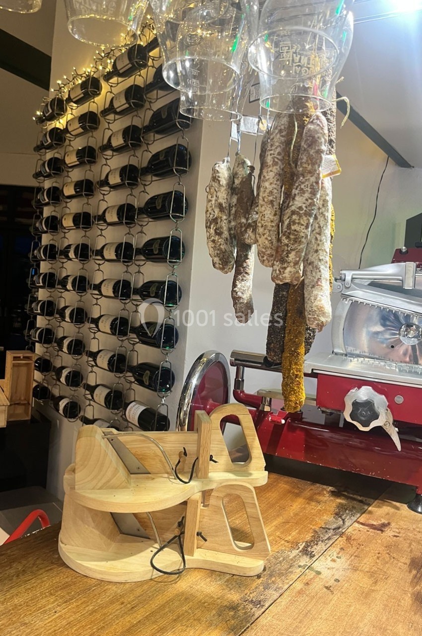 Charcuterie suspendue près d'une trancheuse rouge, avec des bouteilles de vin alignées en arrière-plan.