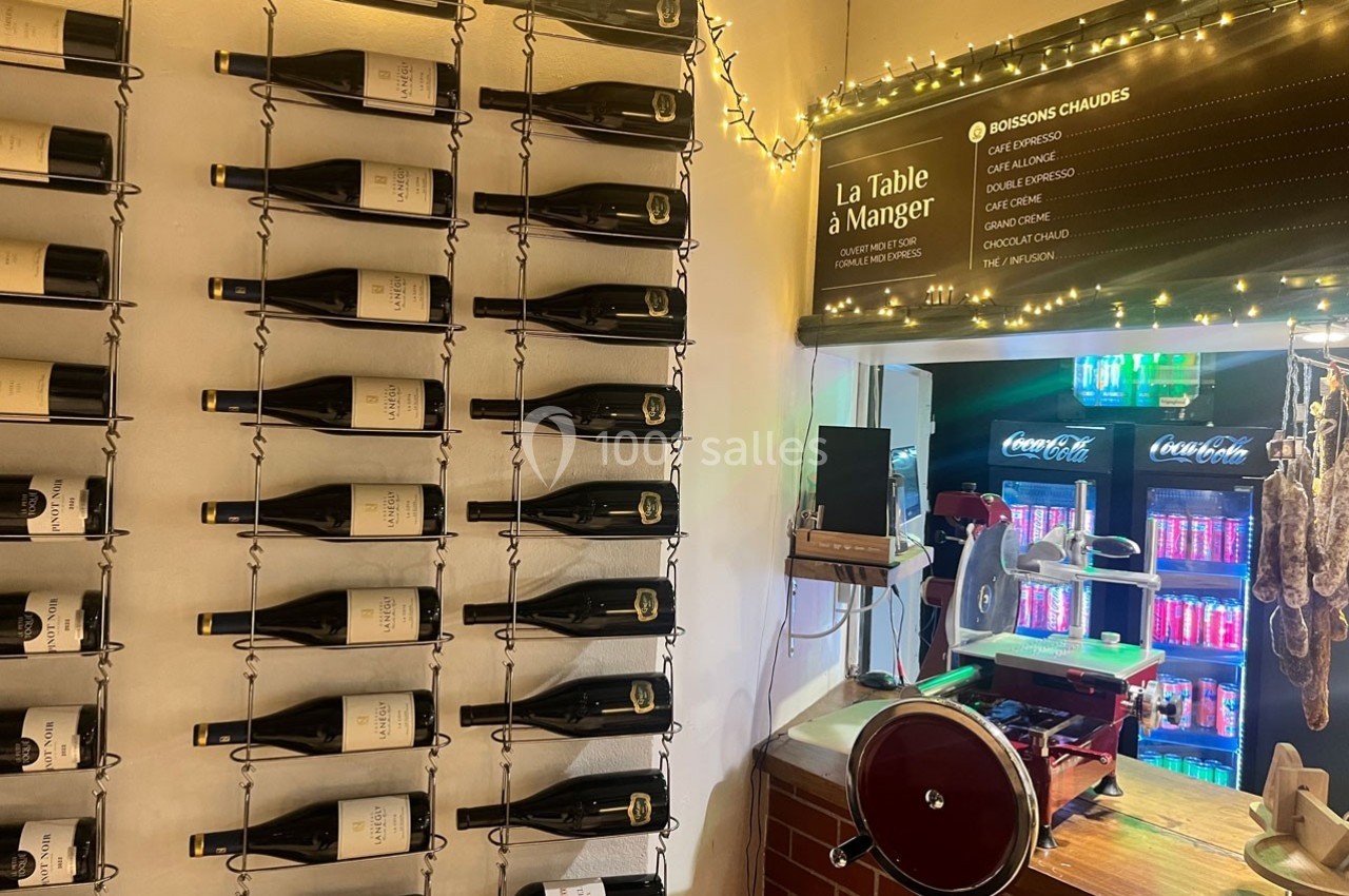 Mur décoratif avec des bouteilles de vin alignées, comptoir avec trancheuse et boissons réfrigérées en arrière-plan.