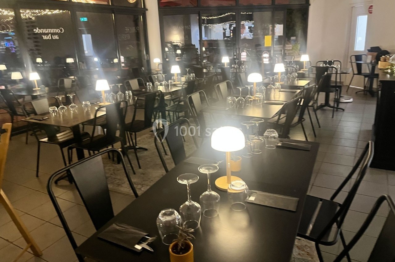 Salle de restaurant avec tables noires dressées, verres et lampes allumées, ambiance chaleureuse et moderne.