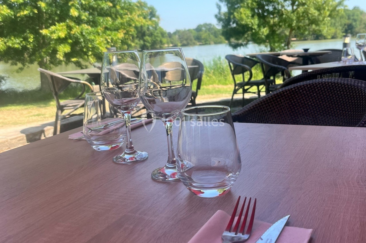 Table dressée en extérieur avec verres et couverts, vue sur un lac et des arbres en arrière-plan.