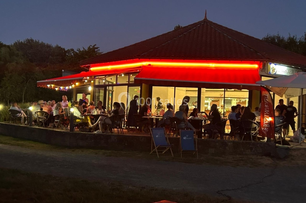 Terrasse d'un restaurant éclairée en soirée, avec des clients attablés sous des lumières et un auvent rouge.