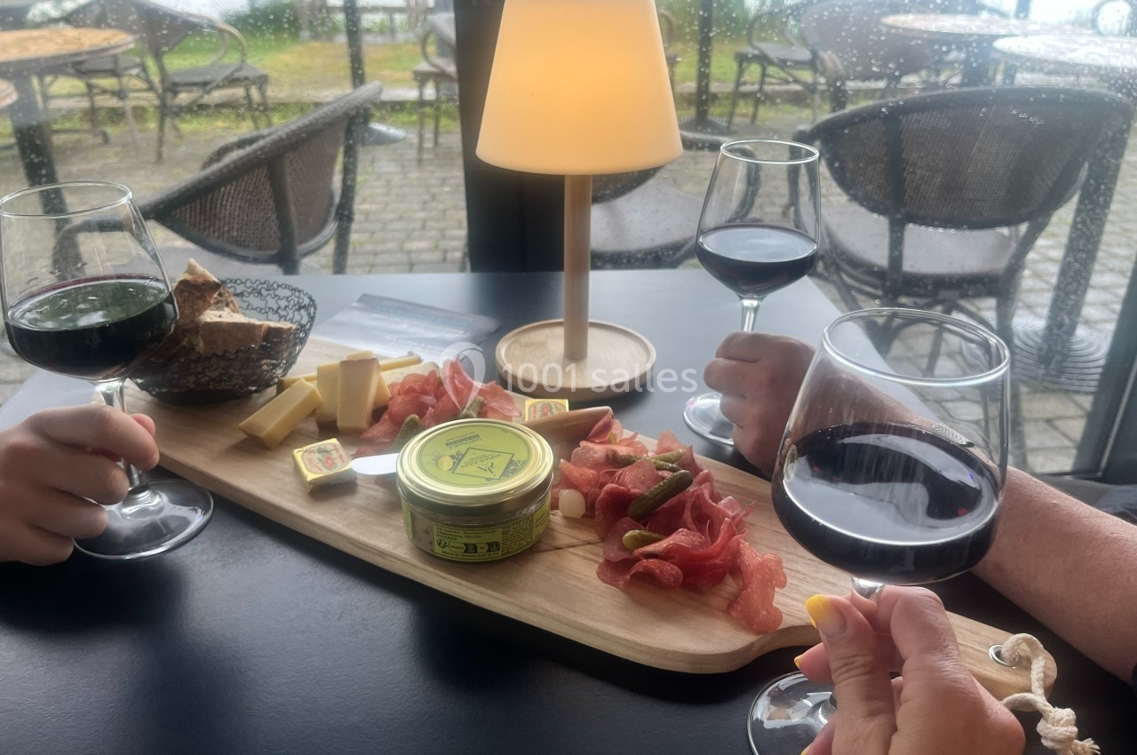 Planche de charcuterie et fromages avec trois verres de vin rouge sur une table, en terrasse par temps pluvieux.
