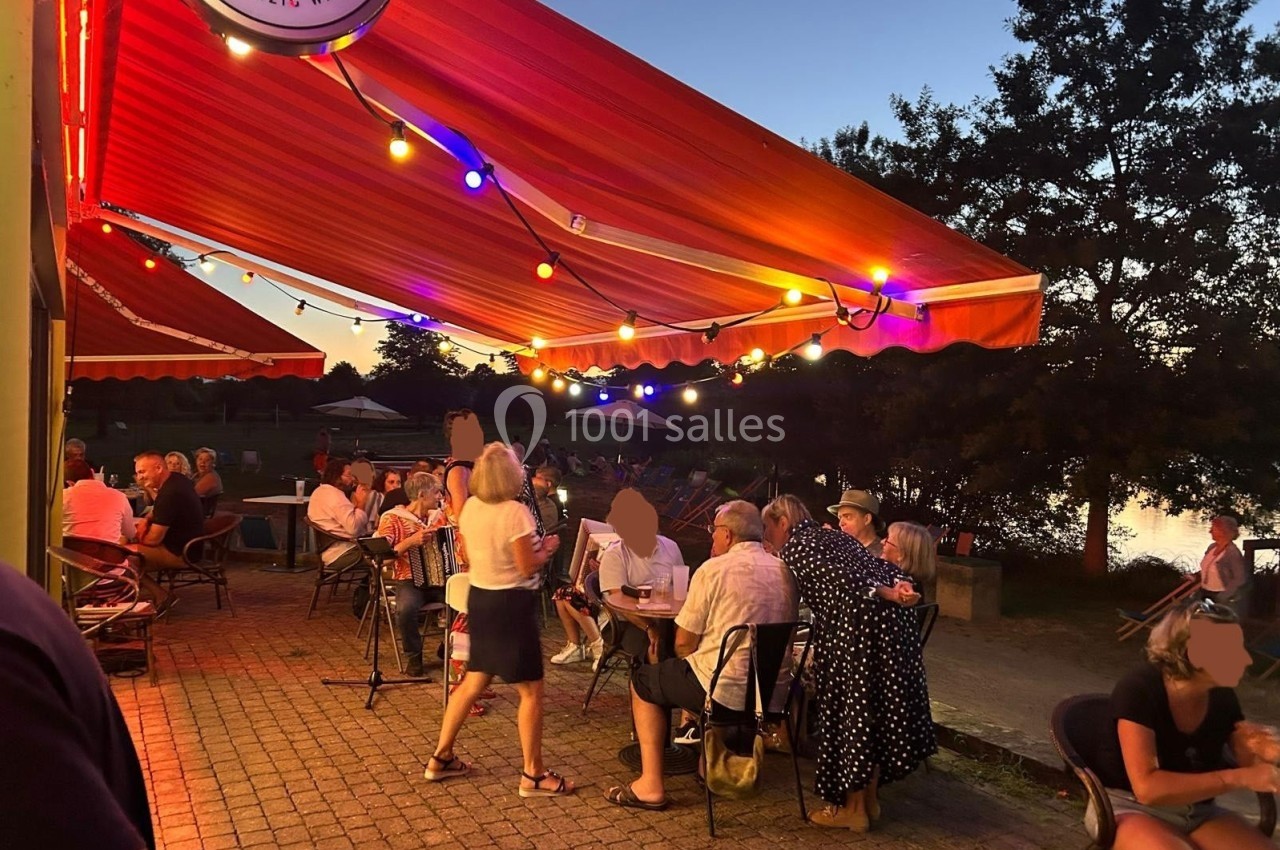 Personnes assises et debout sous des auvents éclairés par des guirlandes lumineuses, en soirée près d'un plan d'eau.