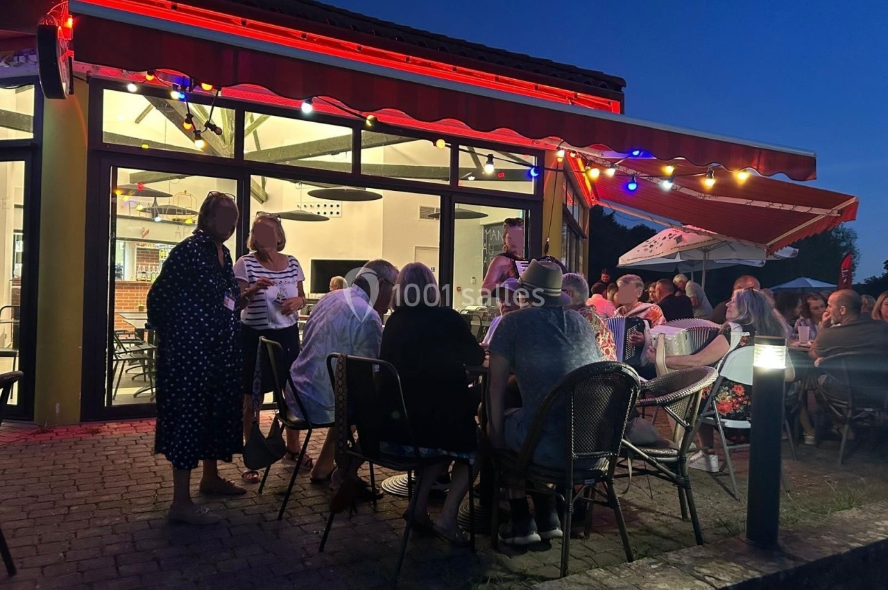 Groupe de personnes assises en terrasse éclairée par des guirlandes lumineuses, devant un bâtiment vitré au crépuscule.