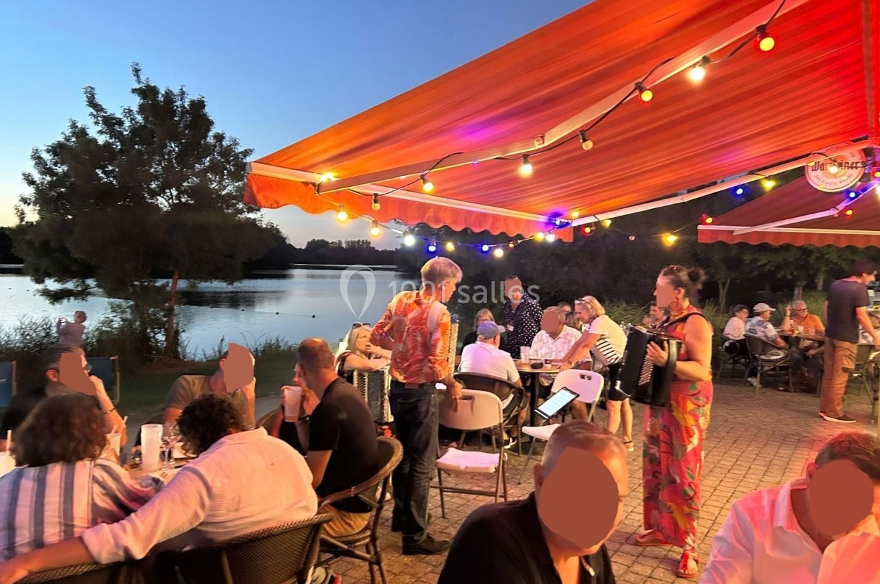 Terrasse animée au bord d'un lac au coucher du soleil, avec des personnes dînant sous des guirlandes lumineuses.