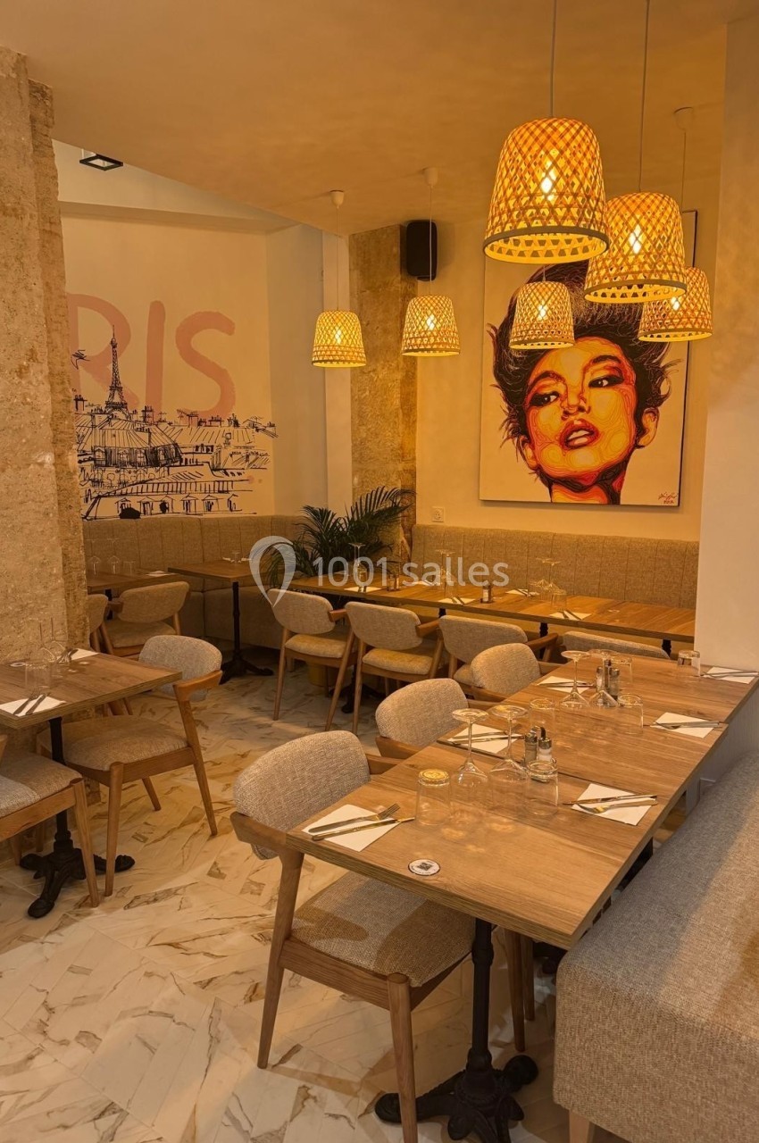 Salle de restaurant chaleureuse avec tables en bois, chaises rembourrées, luminaires suspendus et tableau coloré au mur.