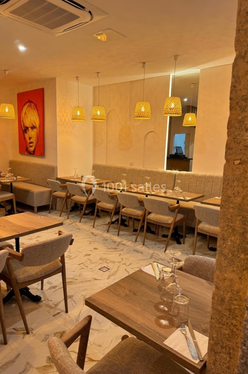 Salle de restaurant chaleureuse avec tables en bois, chaises rembourrées, éclairage suspendu et un tableau coloré au mur.