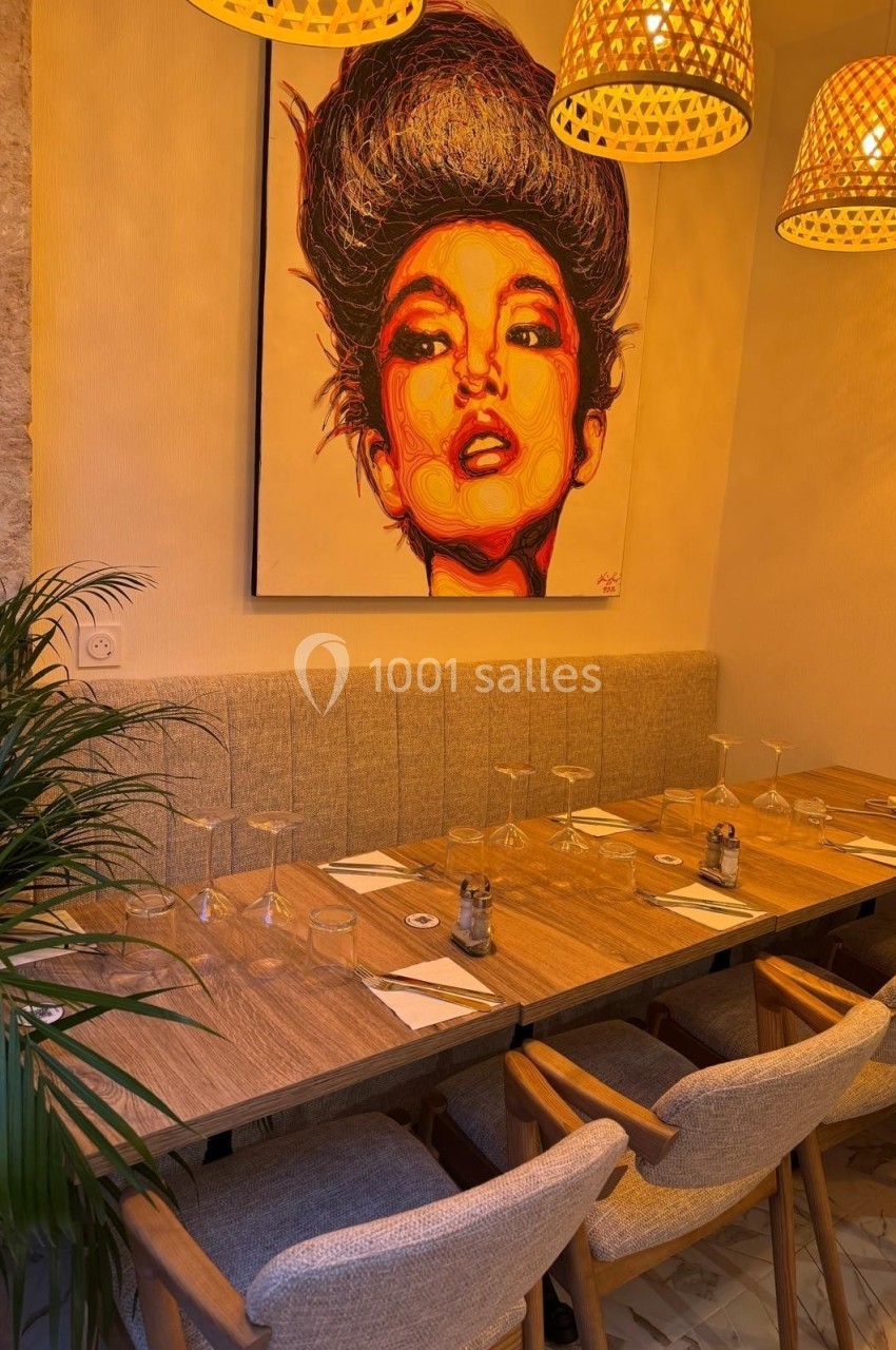 Table en bois dressée pour un repas, entourée de chaises, sous des lampes suspendues et un tableau coloré au mur.