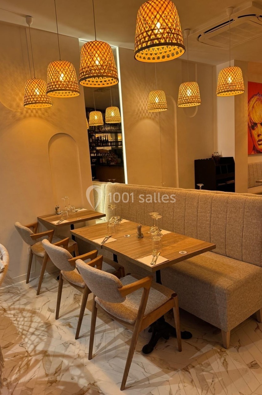 Salle de restaurant chaleureuse avec tables en bois, chaises rembourrées et suspensions en rotin éclairées.