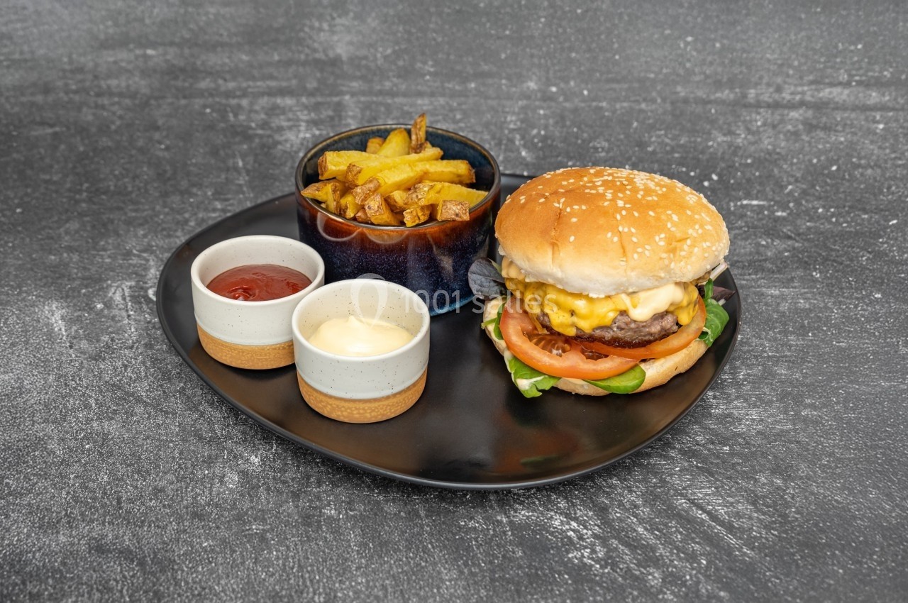 Assiette contenant un burger au fromage, des frites dans un bol et deux sauces servies dans des ramequins.